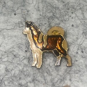 May include: A metal enamel pin shaped like a dog in a standing profile position. The dog features a color palette of dark brown, cream, and gold, with a curled tail and pointed ears, rendered in a glossy finish with defined metallic outlines.