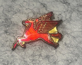 Op de afbeelding: Een rode en gouden emaille pin met een gevleugeld paard, Pegasus, in volle vlucht. De pin heeft een gedetailleerd ontwerp met gouden accenten op de vleugels en het lichaam. Een verzamelobject.