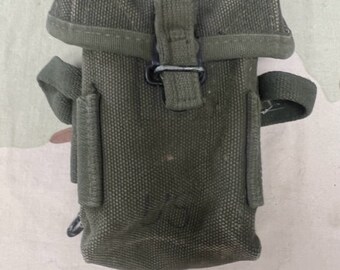 Op de afbeelding: Een groene canvas militaire tas met een klep sluiting en een metalen gesp. De tas heeft een band om te dragen.