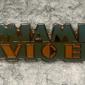 May include: A rectangular enamel pin with the words "MIAMI VICE" in teal and orange. The pin has a gold border and is set against a textured, gray background. The letters are in a bold, stylized font.