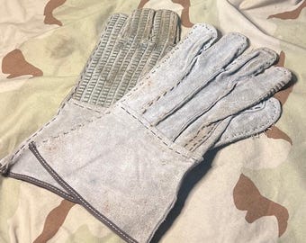 May include: A pair of work gloves made of light gray leather. The gloves have reinforced palms with a textured pattern and are stitched with a contrasting thread. The background is a camouflage pattern.