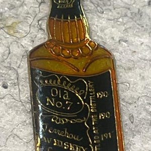 May include: A gold-colored pin in the shape of a whiskey bottle. The pin has a black label with the words "Old No. 7" and "Perrehou Whiskey" in gold lettering. The bottle is outlined in black.