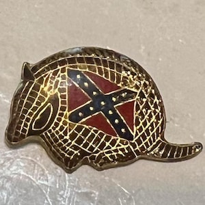 May include: A vintage pin in the shape of an armadillo. The pin features a gold-colored outline with a red, white, and blue emblem at the center. The emblem is a cross design.