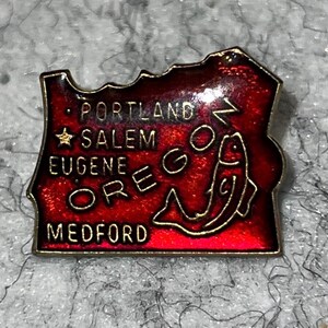 May include: A red and gold enamel pin in the shape of the state of Oregon. The pin features the names of cities Portland, Salem, Eugene, and Medford. A gold fish graphic is on the right side of the pin.
