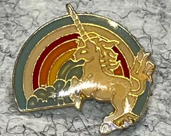 May include: A gold-toned metal enamel pin featuring a prancing unicorn in front of a semi-circular rainbow. The rainbow consists of bands of muted green, red, orange, and yellow. The unicorn is depicted in a light tan color with gold outlines.