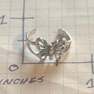 May include: A silver-colored butterfly toe ring. The ring features a detailed butterfly design in the center, with the wings spread open. The ring is approximately 1 inch in length.