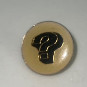 May include: A gold-colored pin with a black question mark on a cream-colored background.