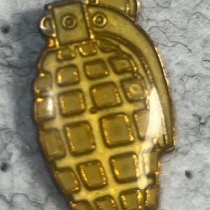 May include: A gold-colored grenade pin with a textured surface. The pin has a detailed design, including a ring and a series of square indentations. The pin is likely made of metal and has a shiny finish.