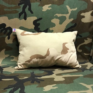 May include: A tan-colored pillow with a camouflage pattern on a camouflage background.