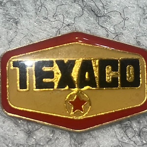 May include: A vintage Texaco pin with a gold background, red border, and black lettering. The pin features the word "TEXACO" and a red star within a gold circle. The pin is a classic piece of automotive memorabilia.