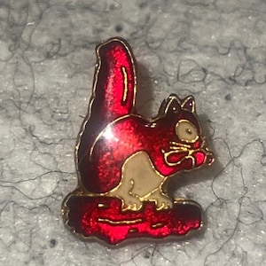 May include: A small metal enamel pin featuring a stylized squirrel with a vibrant red body, a cream-colored belly, and gold-toned outlines. The squirrel is depicted in a seated position with its tail held upright, perched upon a red base.