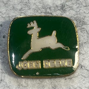 May include: A square, green John Deere pin with a gold border. The pin features a white leaping deer logo and the words "JOHN DEERE" in gold lettering.