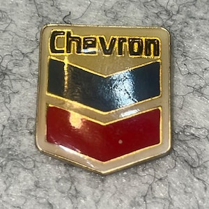 May include: A Chevron pin with a gold border and a shield design. The pin features a gold top section, a blue and black middle section, and a red bottom section. The word "Chevron" is written in black letters.