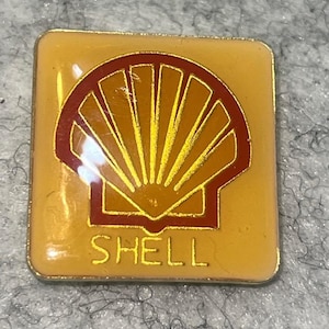 May include: A square, yellow enamel pin featuring the Shell logo. The logo is a red outline of a scallop shell with yellow rays, and the word "SHELL" in gold lettering at the bottom. The pin has a gold border.