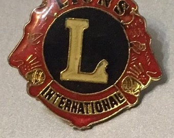 Op de afbeelding: Een rood-gouden Lions International-speld met een zwarte achtergrond en een gouden letter L in het midden. De speld heeft twee leeuwenkoppen aan weerszijden van de letter L.