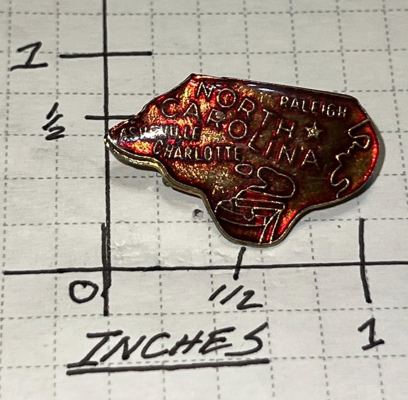 May include: A North Carolina state-shaped pin with the words "North Carolina" and the cities "Asheville", "Charlotte", and "Raleigh" visible. The pin is approximately 1 inch in length and has a red and gold color scheme.