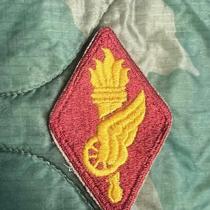 May include: A red diamond-shaped patch with a yellow embroidered design of a torch, a wheel, and a wing.