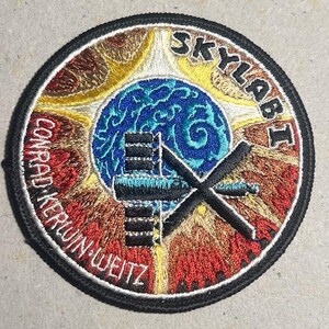 May include: Embroidered Skylab I patch with a black border. The patch features a blue and green globe, a black space station, and the text "SKYLAB I" and "CONRAD KERWIN WEITZ". The background has yellow, brown, and red hues.