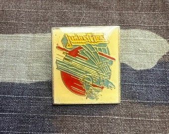 Op de afbeelding: Een gele en blauwe pin met het woord "Judd's Flies" in gele letters. De pin heeft een gestileerd beeld van een vlieg met rode en blauwe vleugels.