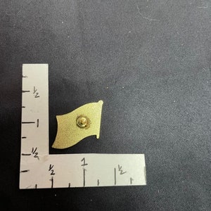 May include: A gold-colored metal pin in the shape of a flag. The flag is waving in the wind. The pin is about 1 inch wide and 1.5 inches tall.