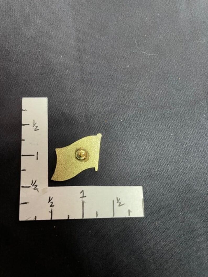 May include: A gold-colored metal pin in the shape of a flag. The flag is waving in the wind. The pin is about 1 inch wide and 1.5 inches tall.