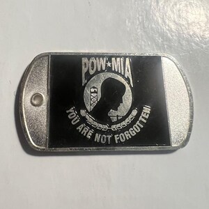 May include: A silver metal dog tag with a black background. The tag features a POW MIA emblem with a silhouette of a soldier's head and the text "You are not forgotten".