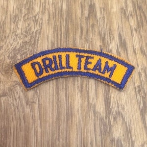 May include: A yellow and blue embroidered patch with the text "DRILL TEAM" in a curved shape.
