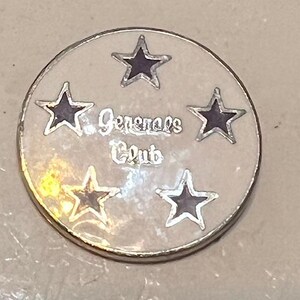 May include: A round, silver-toned pin with a cream-colored center. The pin features five dark purple stars and the words "Generals Club" in a silver script. The pin has a silver border.