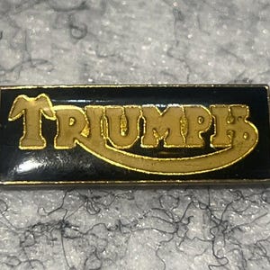 May include: A rectangular pin with the word "TRIUMPH" in gold lettering on a black background. The pin has a gold border and is likely a vintage or collectible item.