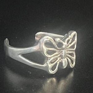 May include: A silver-colored ring featuring a butterfly design. The butterfly has openwork wings and is the focal point of the ring. The ring has a curved band and a delicate, nature-inspired aesthetic.