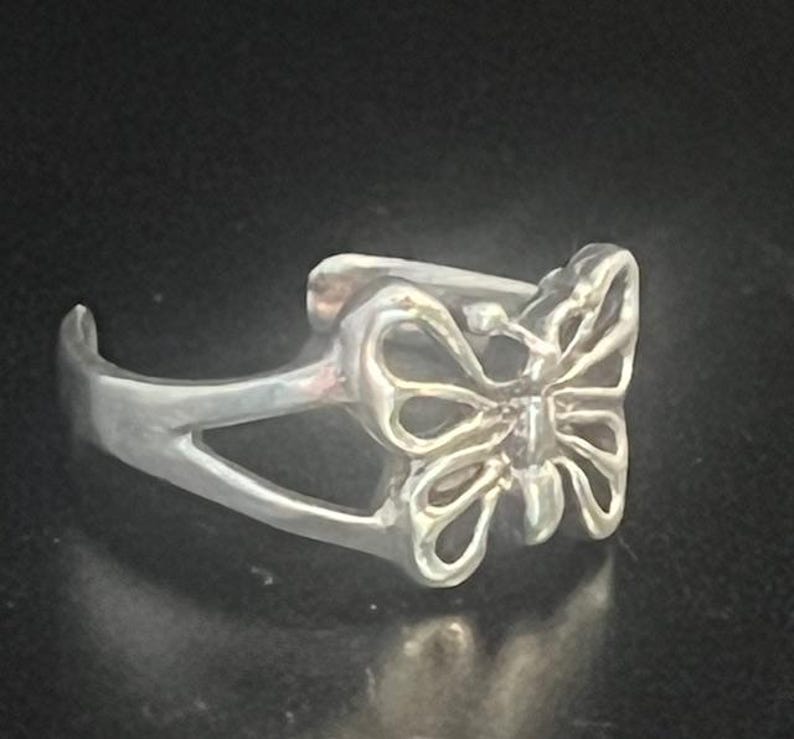 May include: A silver-colored ring featuring a butterfly design. The butterfly has openwork wings and is the focal point of the ring. The ring has a curved band and a delicate, nature-inspired aesthetic.