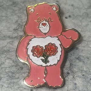 May include: A pink enamel pin shaped like a cartoon bear with a white belly patch featuring two red flowers with smiling faces. The bear has a small red heart on its nose, gold-toned metal outlines, and is depicted in a standing position.