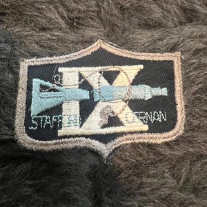 May include: A black and white embroidered patch with a stylized 'X' and a blue and white image of a gun. The text 'STAFFORD' and 'ORRION' are embroidered in white on the patch.