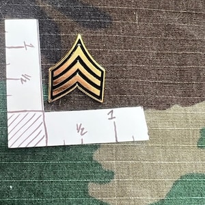 May include: A gold-colored military rank insignia with three black stripes. The insignia is shaped like a chevron and is about 1 inch wide.
