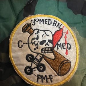 May include: A yellow and white embroidered patch with a skull and crossbones design. The text "3rd MED BN" and "FMF" are visible on the patch.