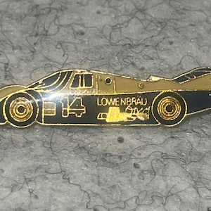 May include: A metal lapel pin shaped like a race car with a black and gold color scheme. The car features the number 14 and the text LOWENBRAU printed on the side. The design includes gold-toned wheels and a streamlined body profile.