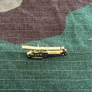 May include: A gold-colored pin of a black helicopter with red and white accents. The helicopter is flying with its blades spinning.