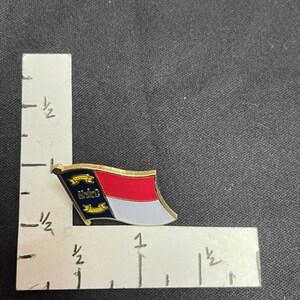 May include: A gold-colored pin with the North Carolina state flag. The flag is red, white, and blue with a yellow star in the blue triangle. The pin is about 1 inch in length.