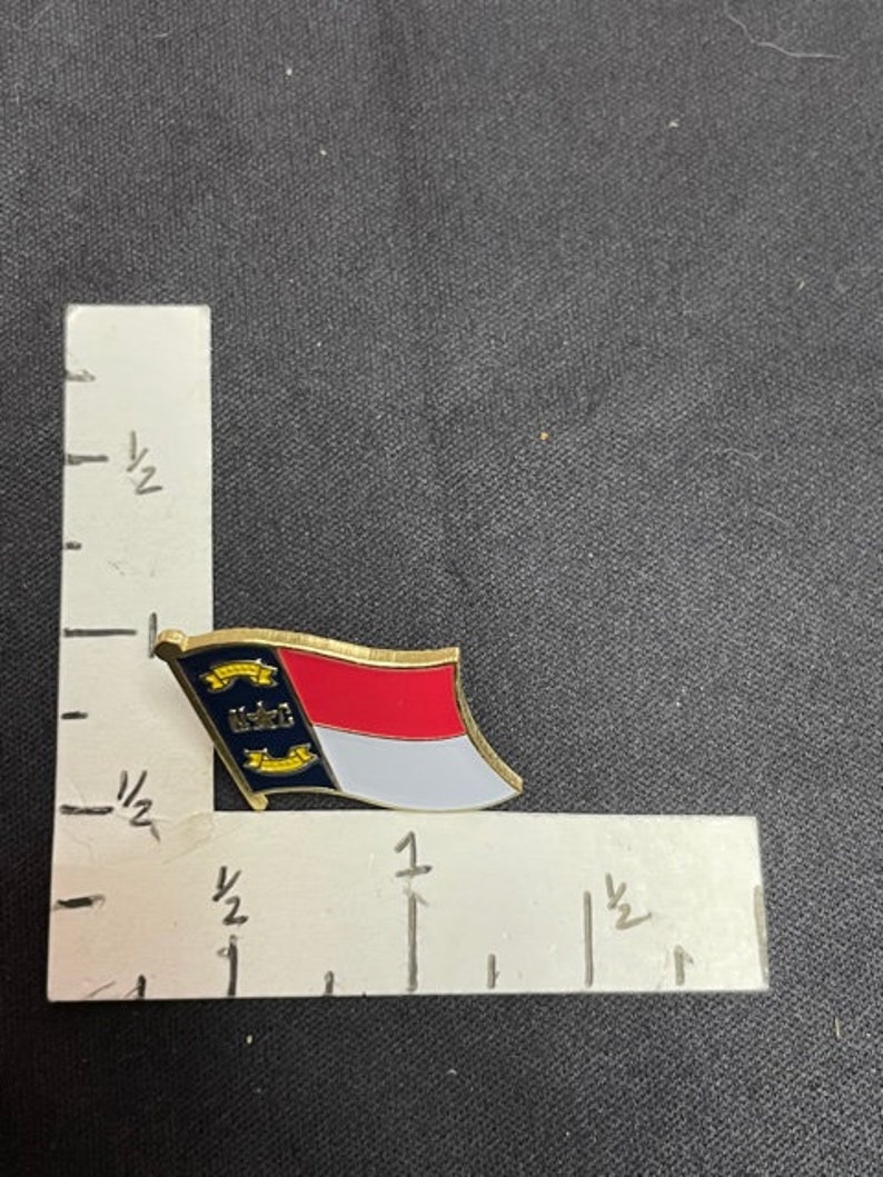 May include: A gold-colored pin with the North Carolina state flag. The flag is red, white, and blue with a yellow star in the blue triangle. The pin is about 1 inch in length.