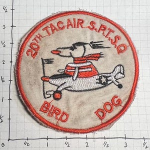 May include: A red, white, and blue embroidered patch with the text "20th TAC AIR S.R.T.S.Q" and "BIRD DOG". A cartoon dog in a red scarf is flying a plane.