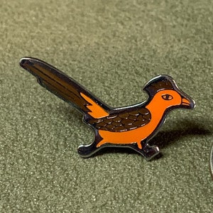 May include: A metal pin of a roadrunner bird with brown and orange colors. The bird is facing right and has a long tail.