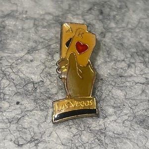May include: A gold-toned Las Vegas souvenir pin featuring a hand holding two playing cards, an Ace and a King of hearts. The pin has the words "Las Vegas" at the base.