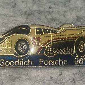 May include: A gold-toned metal enamel pin shaped like a race car, specifically a BFGoodrich Porsche 962. The car features black detailing, the text BFGoodrich on the side, and a blue rectangular base with the text BFGoodrich Porsche 962 printed in white.