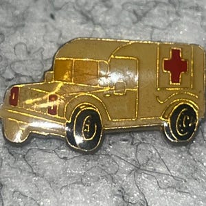 May include: A vintage ambulance pin with a gold-colored outline and a tan body. The pin features a red cross on the side and black wheels. The ambulance pin is a collectible item.