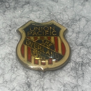 May include: A vintage Union Pacific Railroad pin in a shield shape. The pin features the words "Union Pacific" at the top and "The Overland" across the center. The design includes red and gold accents.
