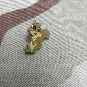 May include: A gold-colored metal pin featuring a cartoon squirrel with red lips and brown fur. The squirrel is holding its tail in its paws.