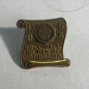 May include: A gold-colored metal pin with a scroll design. The scroll has the text "Roll Call for Men of Distinction" and the year "1965". The pin features a blue and gold emblem with the text "American Legion".