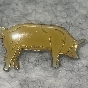 May include: A gold-toned metal enamel pin shaped like a pig in profile. The pig is colored in a light tan or beige shade with thin, raised gold-colored outlines defining its body, legs, snout, and curled tail. The pin has a smooth finish.
