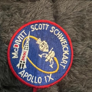 May include: A blue and yellow embroidered patch with the text "McDivitt Scott Schweickart Apollo IX" and a depiction of a spacecraft in orbit around the Earth.