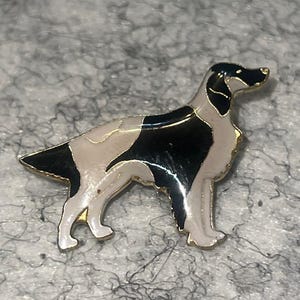 May include: A dog-shaped enamel pin in black, white, and gold. The pin features a detailed design of a dog with a black head and tail, white body, and gold trim. The dog is standing in a side profile.